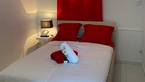 A neatly made bed with white bedding, red pillows, and a red rose. There is a small bedside table with a lamp and a decorative item. - Le Lodge Phoenix, F2 in residence with swimming pool (Sainte-Rose)