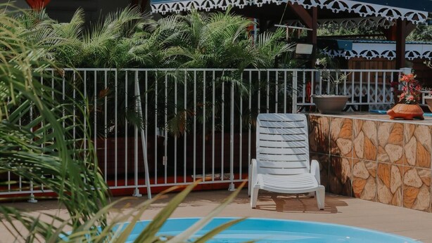 A pool area with a white chair, a stone wall, and a metal fence. - Le Lodge Phoenix, F2 in residence with swimming pool (Sainte-Rose)