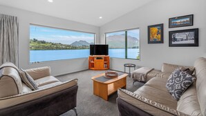 A spacious living room with a large window offering a scenic view of a lake and mountains. There is a leather sofa, a wooden coffee table, and a television on a wooden stand. - Frankton House - Queenstown Holiday Home (Queenstown)