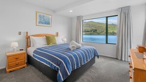A bedroom with a large bed, a nightstand, a dresser, a window with a view of hills and water, and a painting on the wall. - Frankton House - Queenstown Holiday Home (Queenstown)