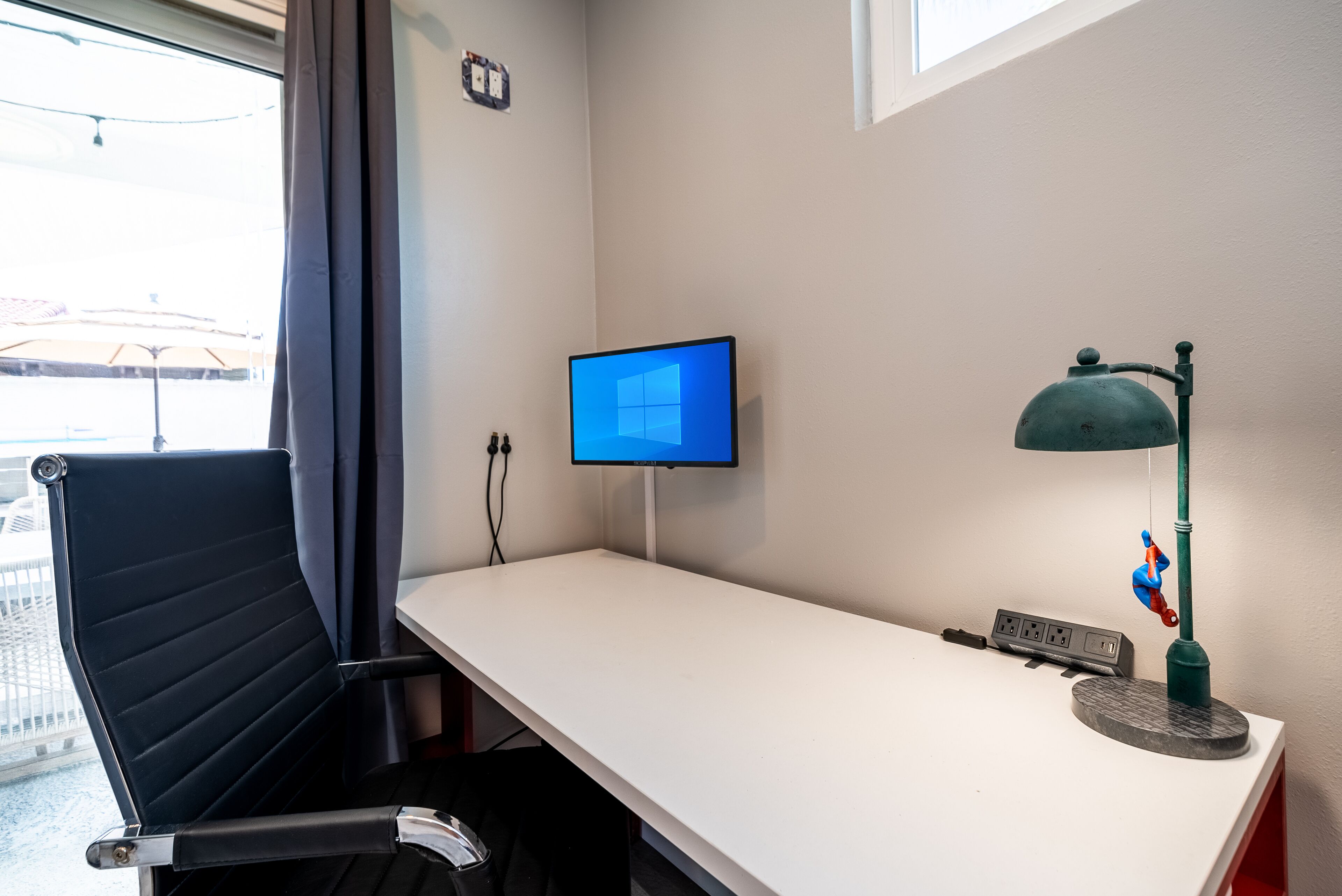 Ein moderner Arbeitsbereich mit einem weißen Schreibtisch, einem schwarzen Bürostuhl, einem Computerbildschirm, einer Schreibtischlampe und einer Steckleiste.