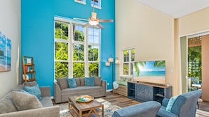 Ein Wohnzimmer mit einem großen Fernseher, grauen Sofas, einem Holztisch und einem Deckenventilator.