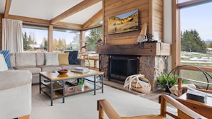 A cozy living room with a fireplace, a sofa, a coffee table, and large windows offering a view of the outdoors.