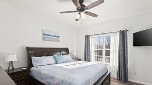 A bedroom with a bed, nightstands, a ceiling fan, a window with curtains, and a wall-mounted TV.