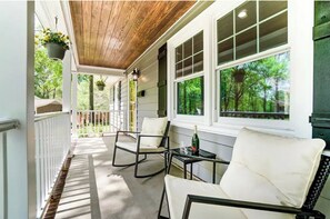 A covered porch with white railings, two rocking chairs, a table with a bottle and glass, and hanging flower baskets.