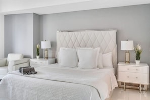 A modern bedroom with a large bed, two bedside tables with lamps, a nightstand, a chair, and a potted plant.