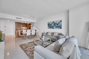 A modern living room with a sofa, coffee table, and a dining area with chairs and a kitchen in the background.
