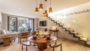 Un espace repas moderne avec une table ronde en bois, des chaises et une table préparée pour le petit-déjeuner. Un escalier avec une rampe en verre est visible à l’arrière-plan.