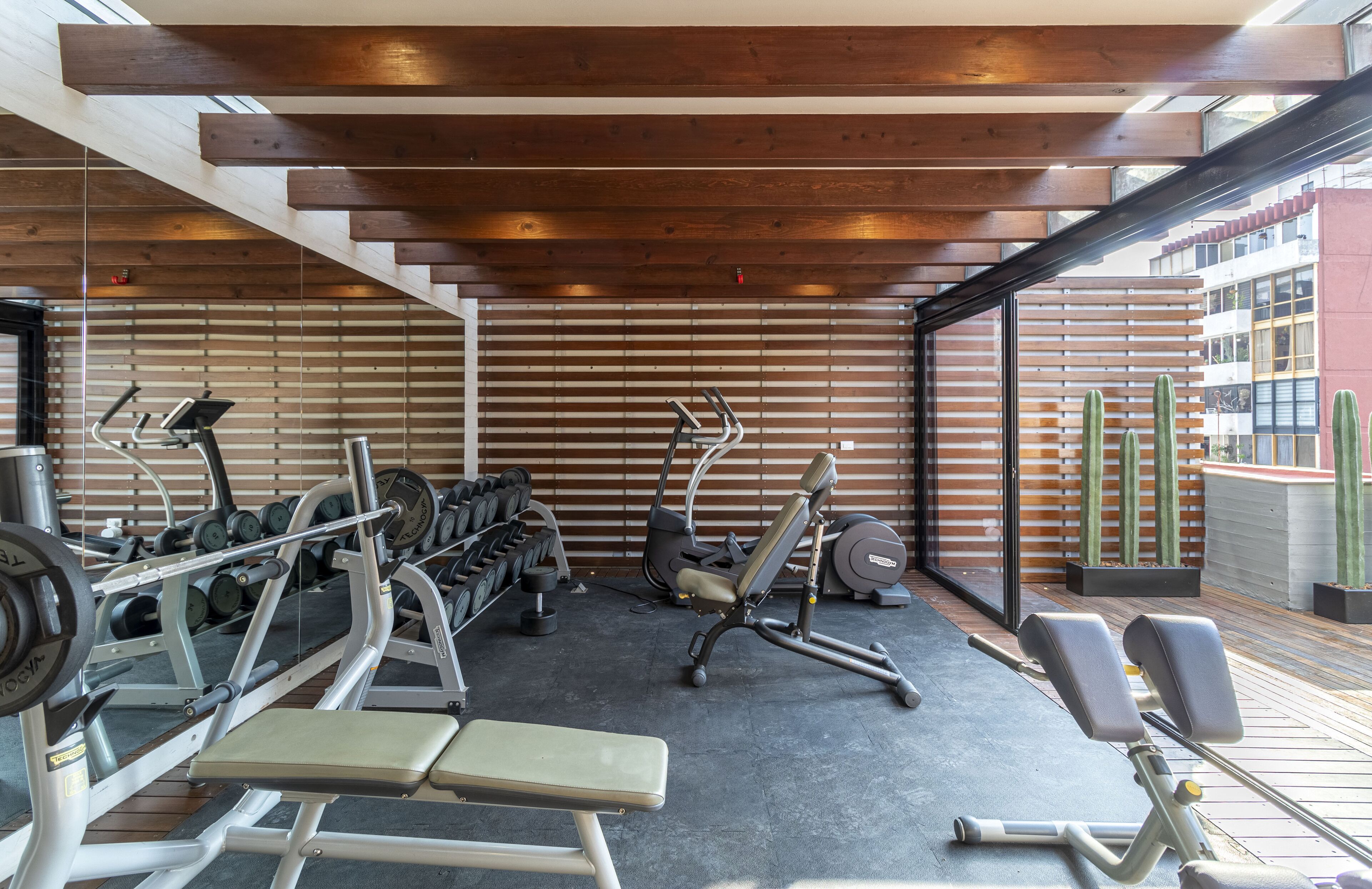 A gym with various exercise equipment, a bench, and a view of the city through large windows.