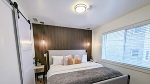 A modern bedroom with a large bed, a wooden panel wall, a window with blinds, and a ceiling light fixture.
