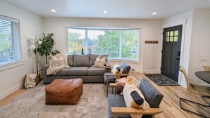 A modern living room with a grey sofa, a brown ottoman, and a blue sofa with patterned cushions. There are large windows with blinds, a black door, and a plant in the corner.