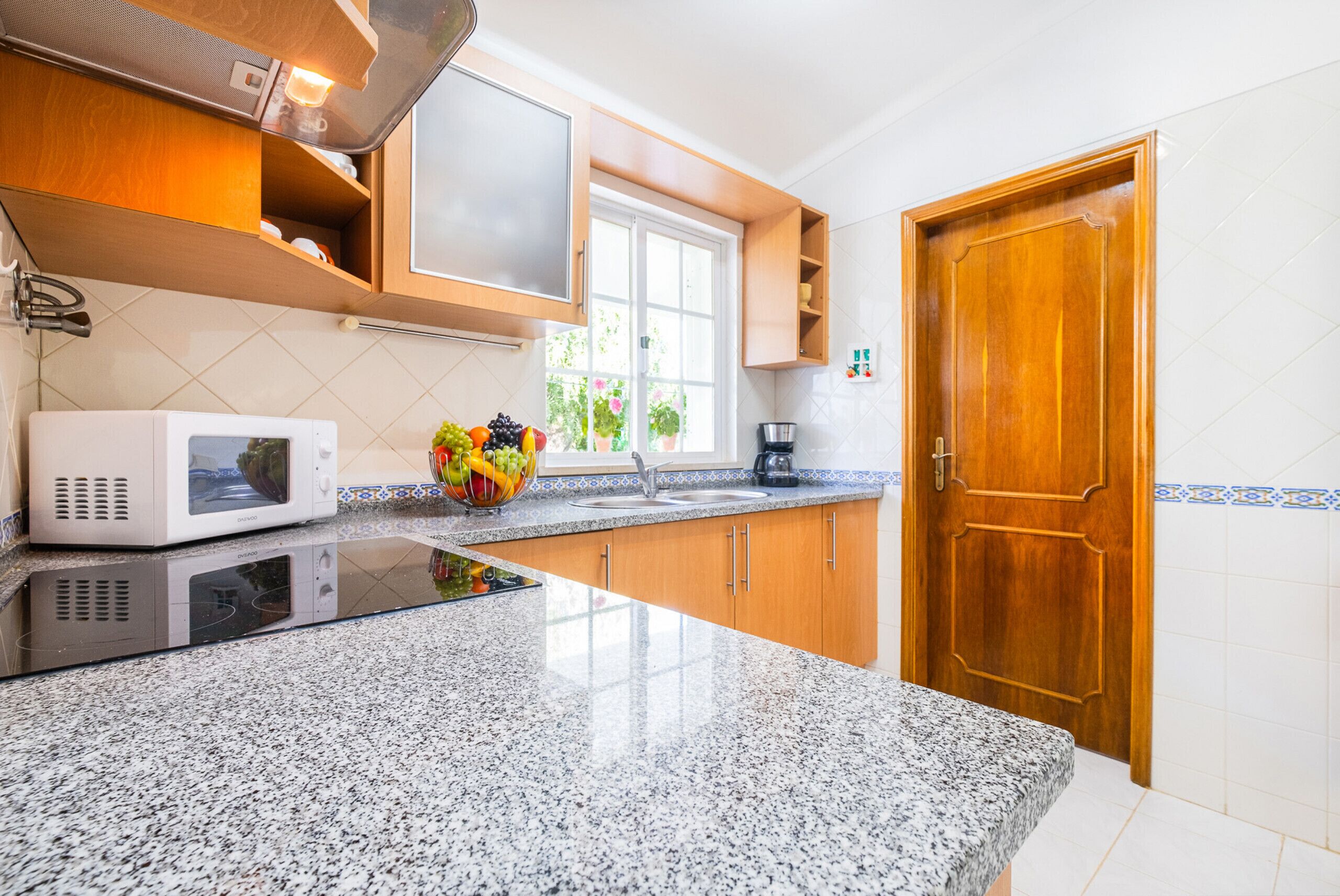 A kitchen with granite countertops, a microwave, a fruit bowl, wooden cabinets, a window, and a wooden door.