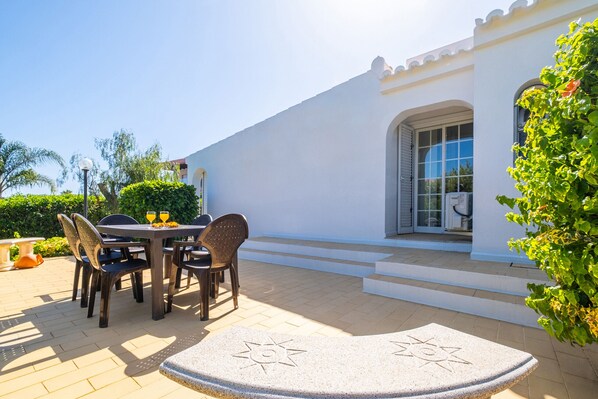 An outdoor patio with a round table and chairs, a stone bench, and a white building with a window and door. - Peaceful villa with pool and barbecue (Armação de Pêra)