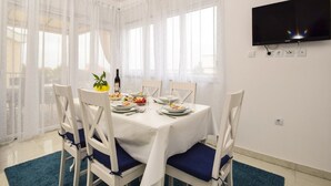 A dining area with a table set for four, white chairs with blue cushions, a TV mounted on the wall, and sheer curtains on the windows. - Apartment in Zaton (Zadar) with Seaview, Balcony, Air condition, WIFI (685-2) (Nin)