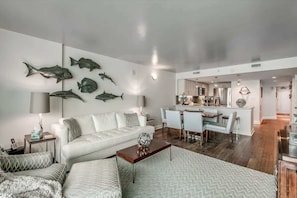 Un salon moderne avec un canapé blanc, une table basse et un coin repas, la cuisine étant visible au fond.