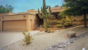 Un paysage désertique avec un grand cactus, un bâtiment doté d’un toit recouvert de tuiles rouges et un arbre.