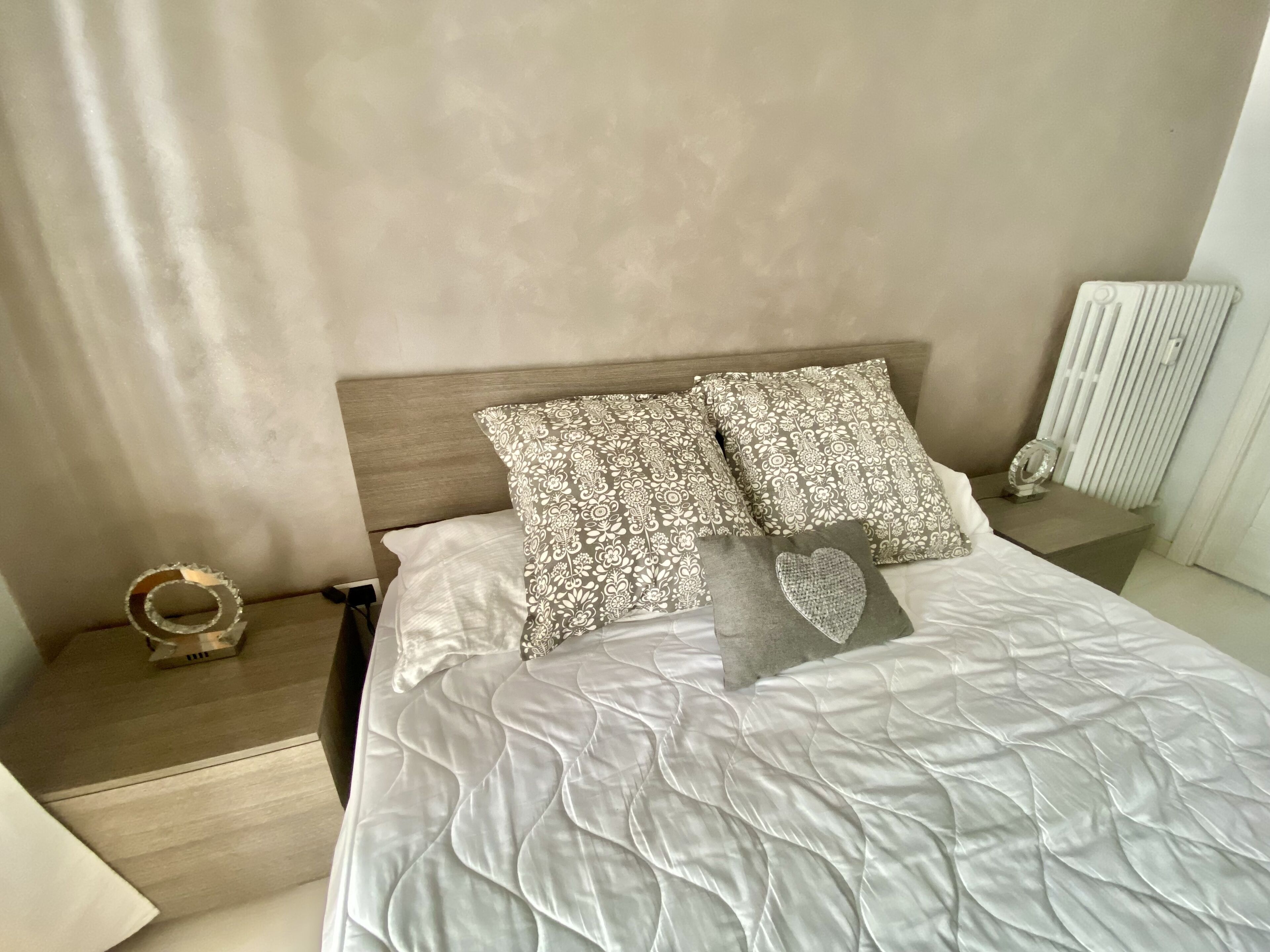 Ein ordentlich bezogenes Bett mit einer gesteppten weißen Decke, zwei gemusterten Kissen und einem herzförmigen Zierkissen. Ein Nachttisch mit einem dekorativen Gegenstand und ein kleiner Tisch mit einer Vase sind zu sehen.