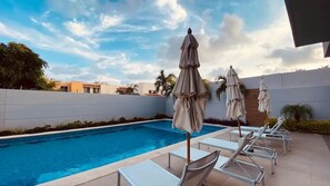Piscina con sillas de descanso y sombrillas, rodeada por un muro bajo y palmeras.