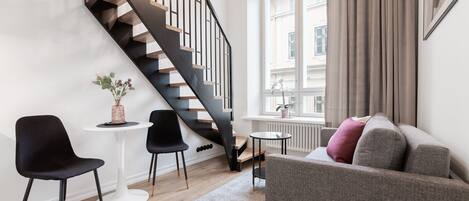 A modern living room with a staircase, a grey sofa, black chairs, a small table, and a window with curtains.