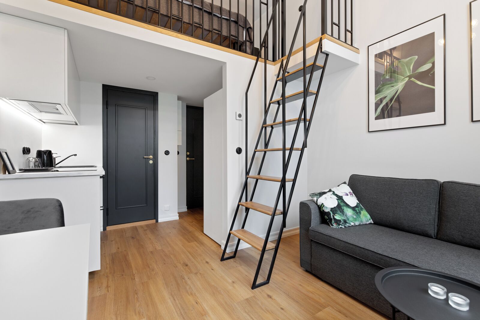 Un apartamento moderno con cocina, un sofá gris, una escalera y una planta enmarcada en la pared.