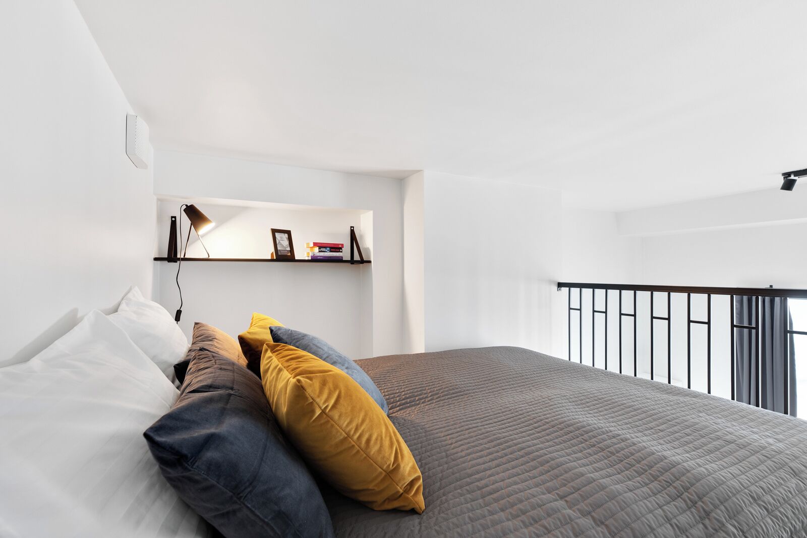 A modern bedroom with a bed, pillows, a shelf with books, and a balcony railing.