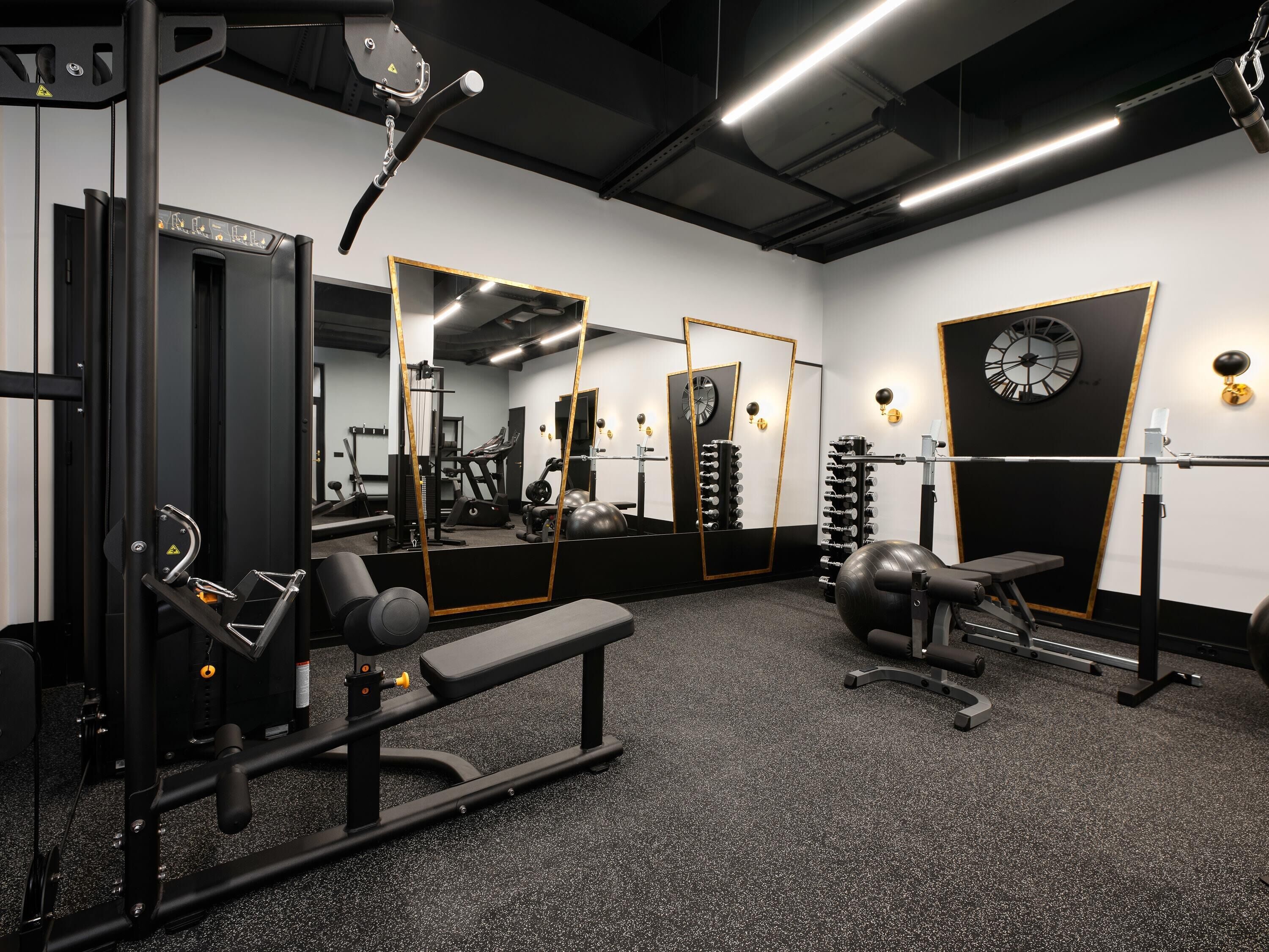 Ein Fitnessstudio mit verschiedenen Trainingsgeräten, Spiegeln und einer Uhr an der Wand.