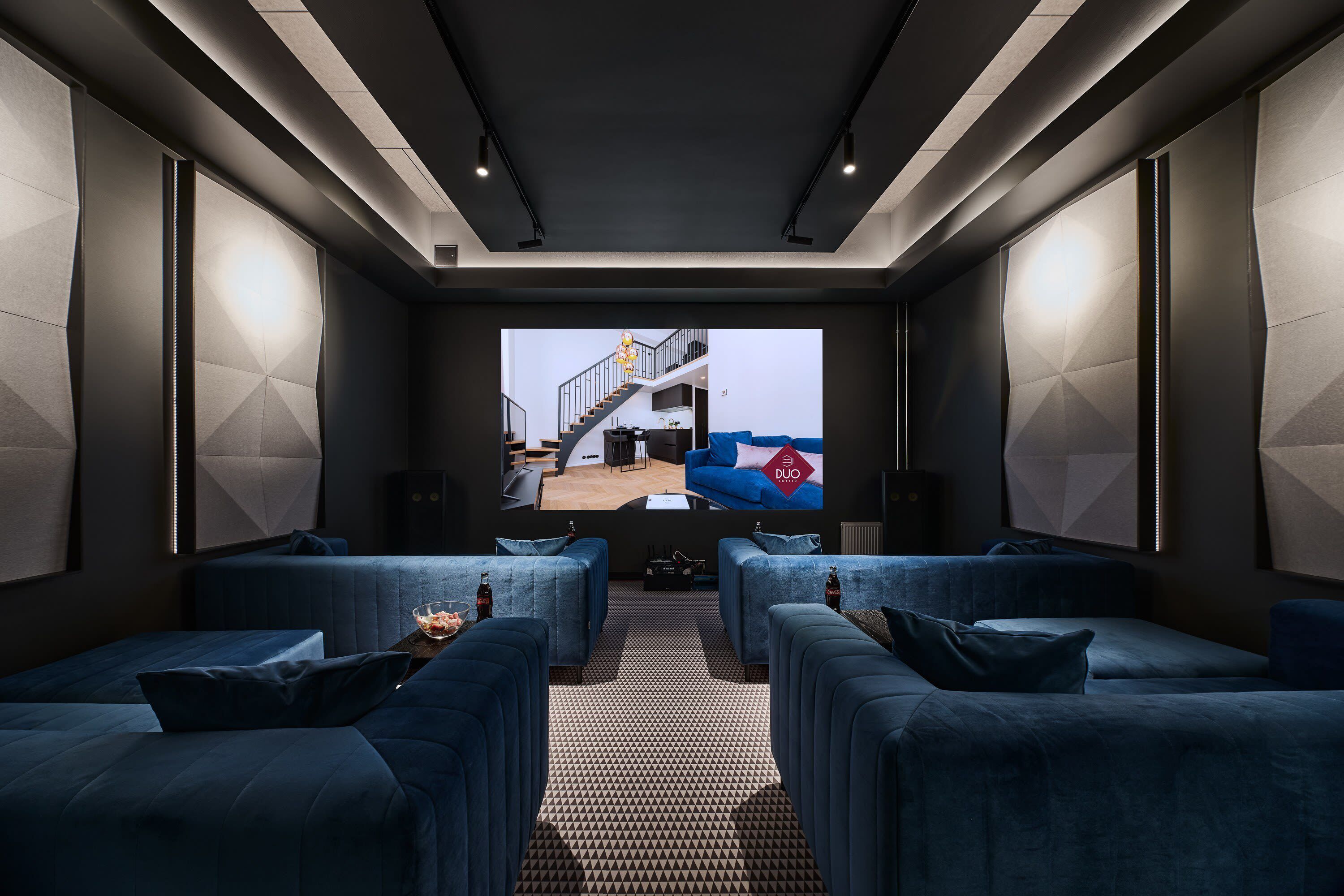 A modern, dark-themed living room with a large screen displaying a room. There are plush blue sofas and a patterned carpet.