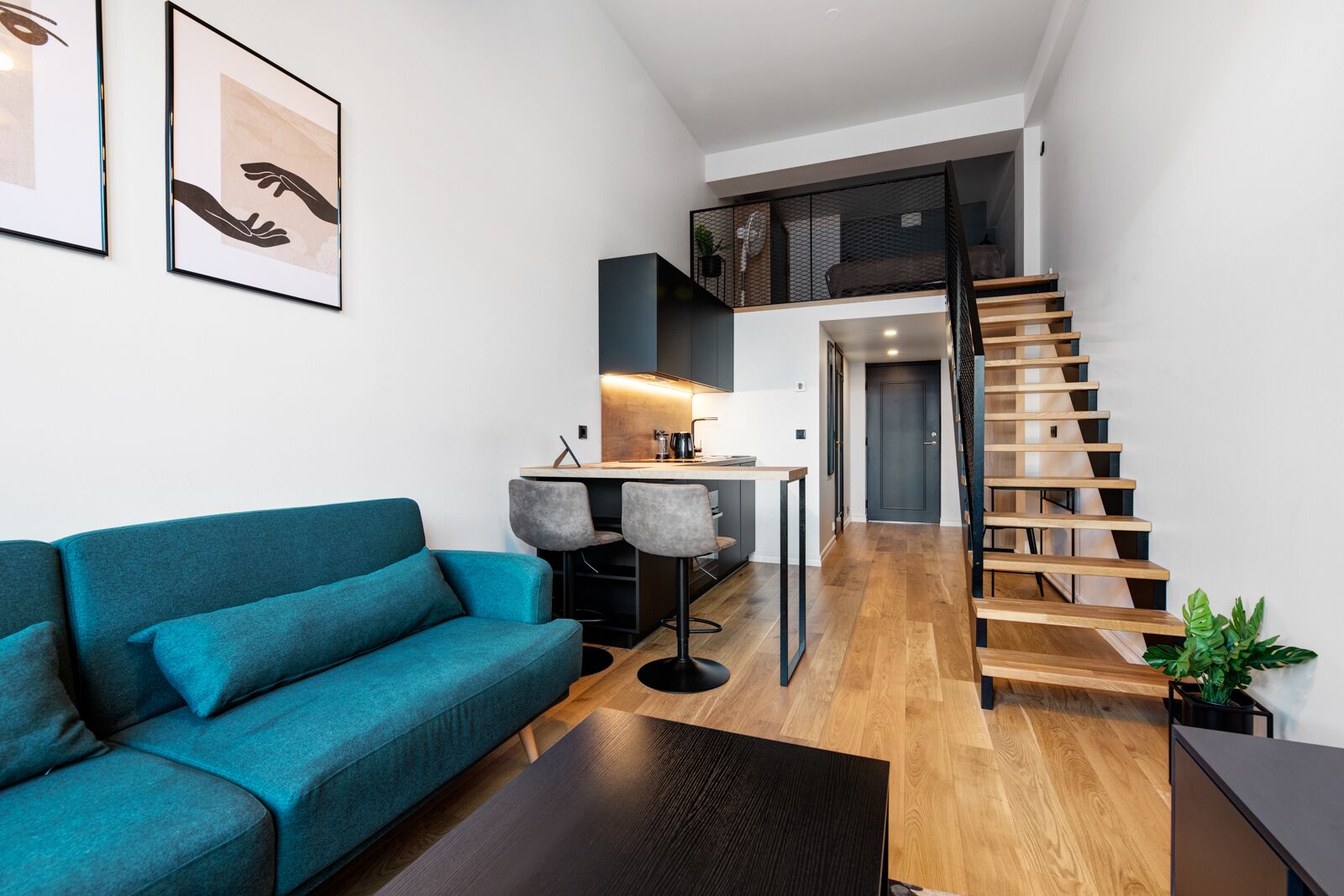 Una sala moderna con un sofá color turquesa, una pequeña zona de cocina y una escalera de madera.
