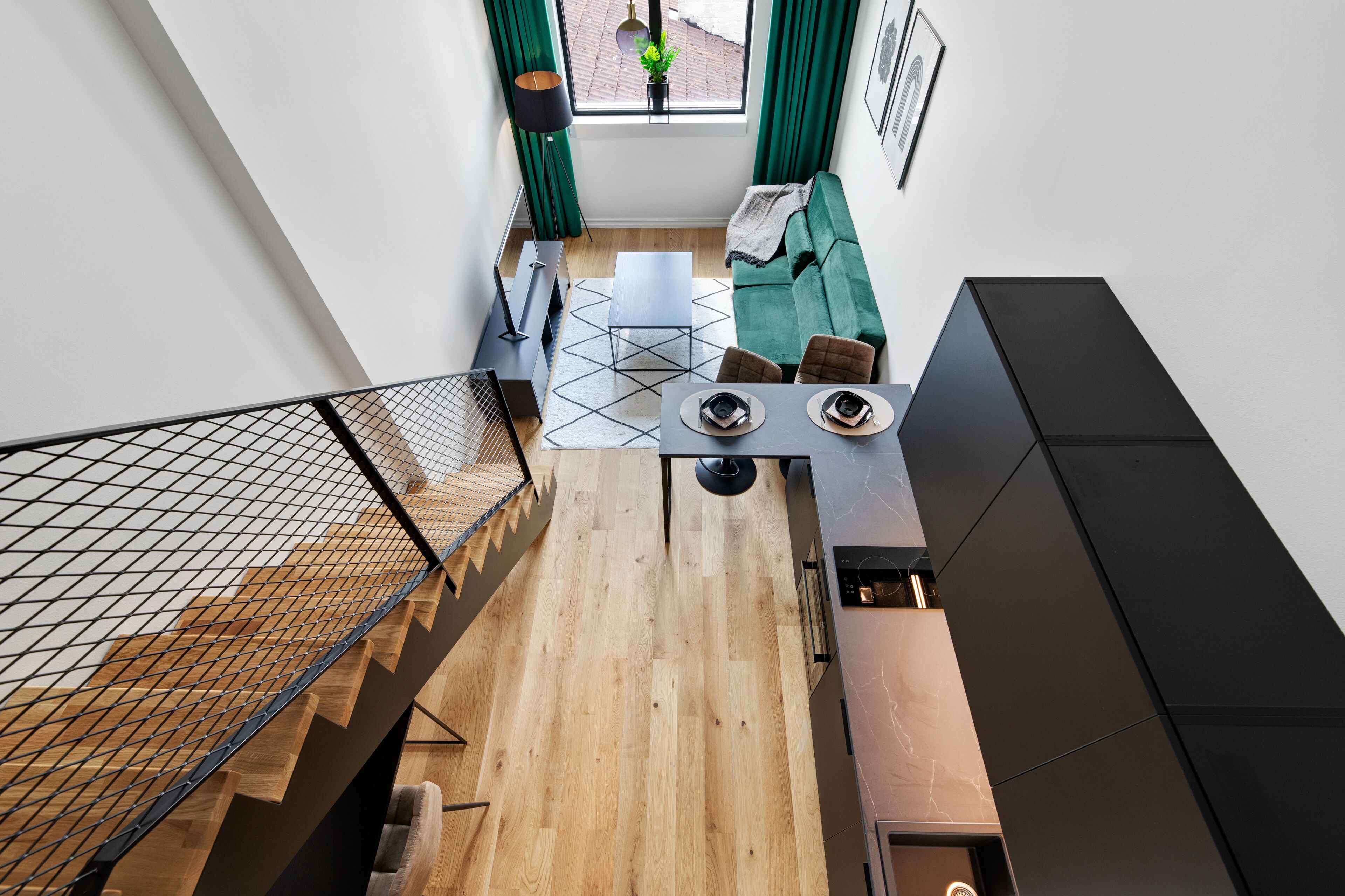 Ein modernes Interieur mit Holzboden, einem grünen Sofa, einem Esstisch mit Stühlen und einer Treppe mit Metallgeländer.
