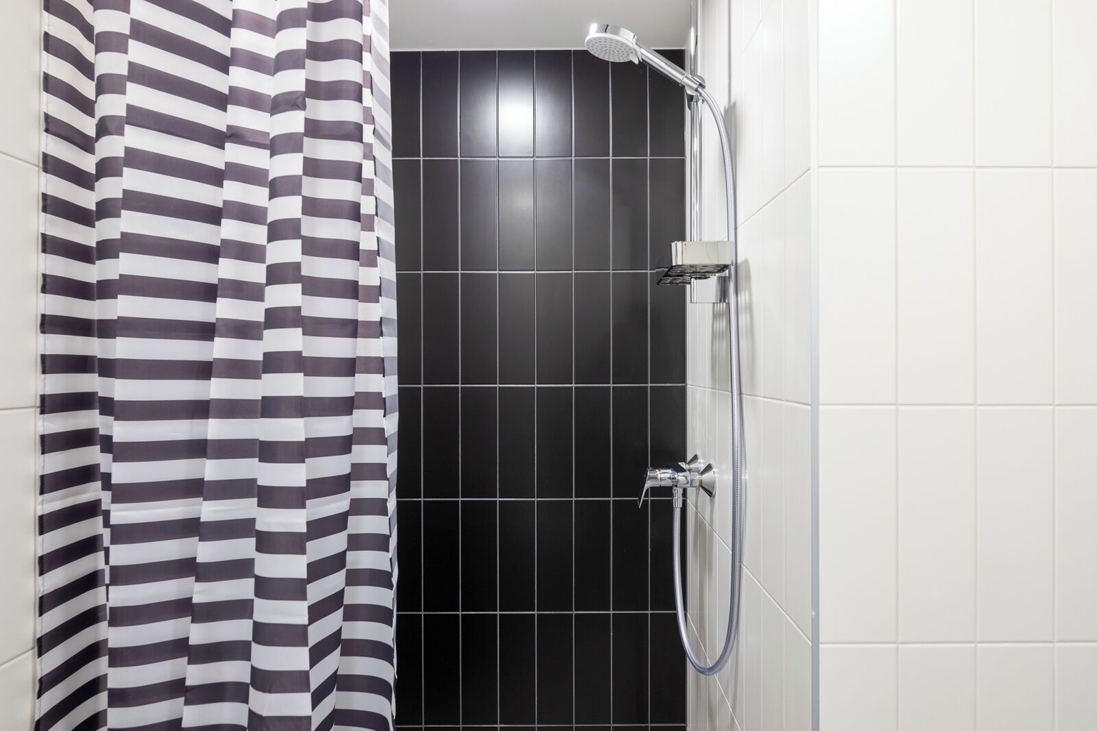 Zona de ducha con pared de azulejos negros, cortina de baño a rayas y ducha de mano.