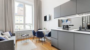 Una cocina moderna con armarios grises, encimera blanca y horno integrado. Hay una zona de comedor con mesa y sillas, y una ventana con cortinas.