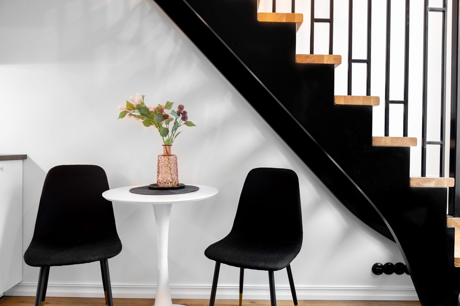 Ein modernes Zimmer mit einem weißen runden Tisch, zwei schwarzen Stühlen, einer Vase mit Blumen und einer schwarzen Treppe mit Holzelementen.