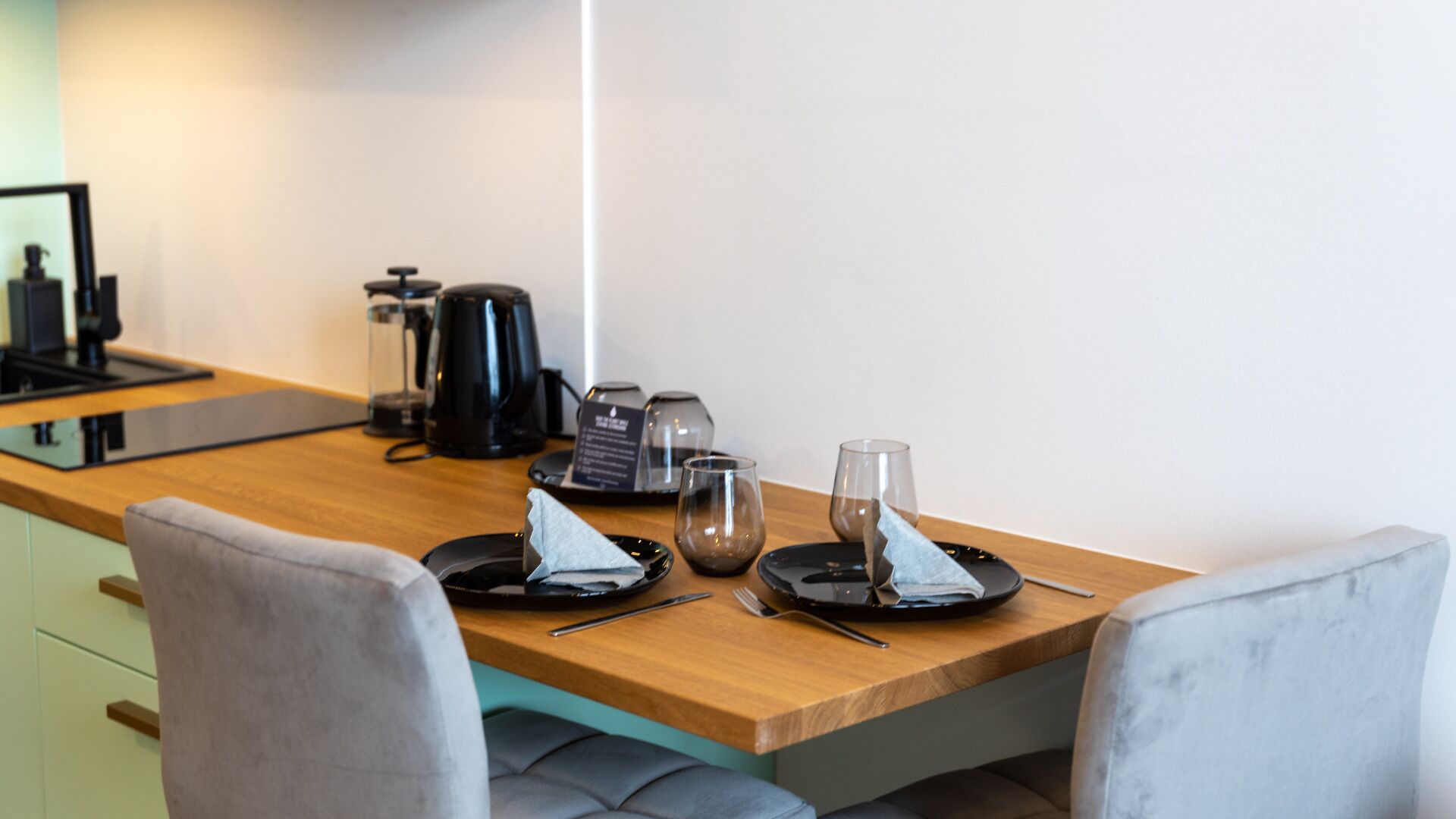 A modern kitchen with a wooden countertop, a coffee maker, and a kettle. There are two chairs and a small table with a napkin and a glass.