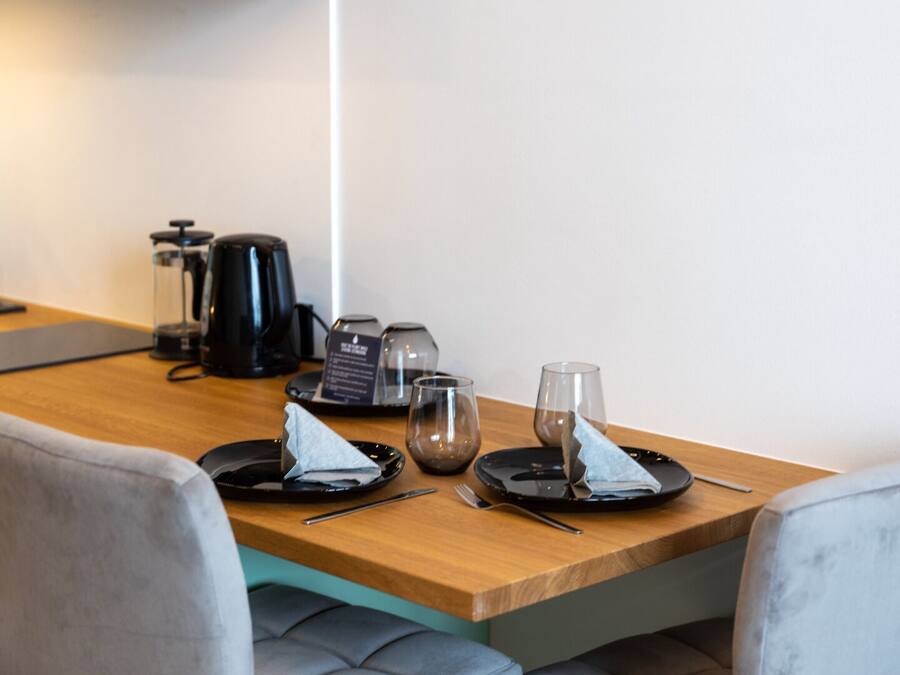 A modern kitchen with a wooden countertop, a coffee maker, and a kettle. There are two chairs and a small table with a napkin and a glass.