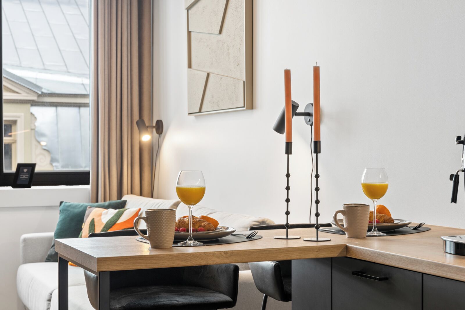 A modern dining area with a wooden table, two glasses of orange juice, a croissant, and a plate of fruit. There is a black chair and a couch with a cushion in the background.