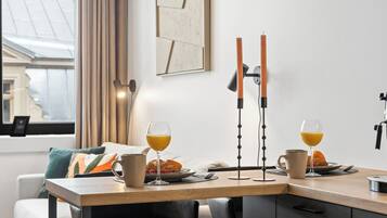A modern dining area with a wooden table, two glasses of orange juice, a croissant, and a plate of fruit. There is a black chair and a couch with a cushion in the background.
