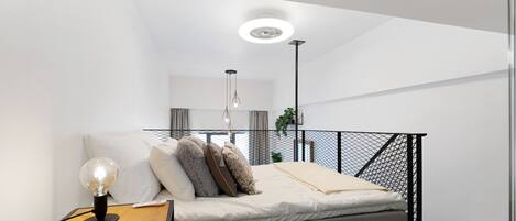 A modern bedroom with a bed, a bedside table with a lamp, a hanging light fixture, and a view of the living area through a balcony.