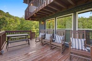 Una terraza de madera con mesa y sillas, un balcón con barandillas y grandes ventanales con vistas a un bosque.