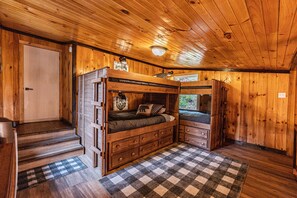 Interior de una cabaña de madera con una litera, un aparador y una alfombra a cuadros.