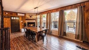 A dining area with a wooden table and chairs, a brick fireplace, and large windows with curtains.