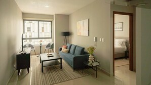 A modern living room with a sofa, coffee table, and a dining area with chairs. There is a large window providing natural light, a floor lamp, and a small table with a vase. - 1BR|1BA Apartment W/ pool, gym, high speed WiFi, located just a 3-minute walk from the new U.S. Embassy (Ciudad de México)