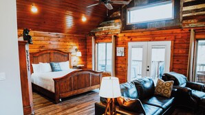 A spacious room with a wooden ceiling, a bed with a headboard, a leather sofa, a small table with a lamp, and a ceiling fan. - Firefly Treehouse - Starlight Haven (Hot Springs)