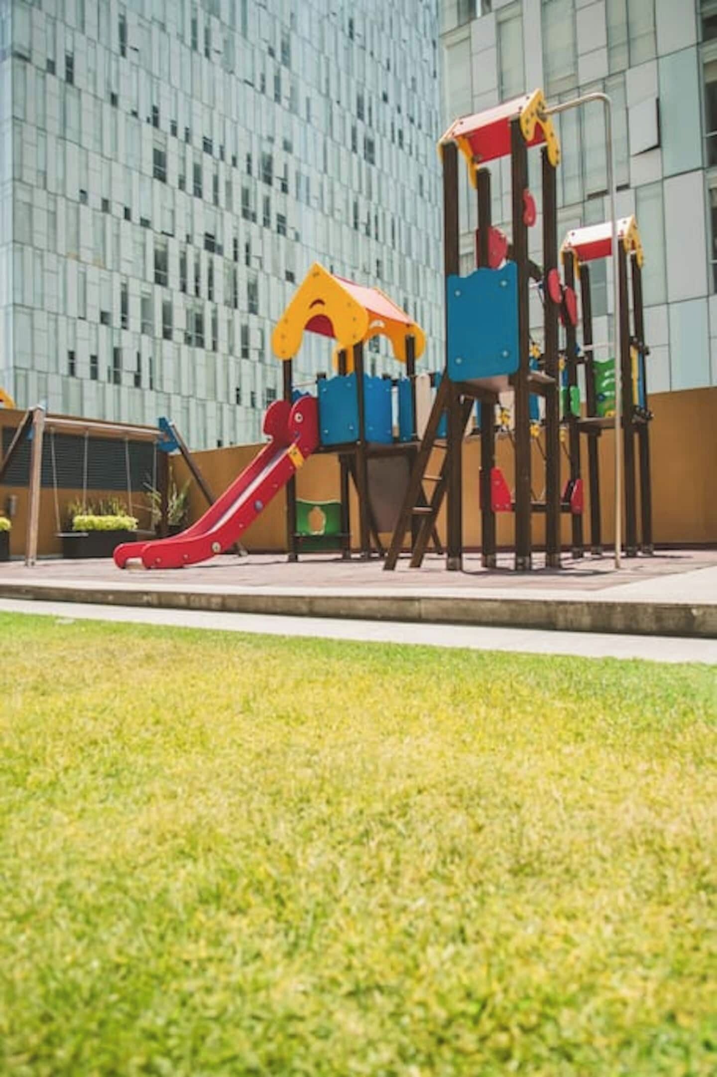 Ein Spielplatz mit einer roten Rutsche und Klettergerüsten vor einem modernen Gebäude mit einer gitterförmigen Fassade.