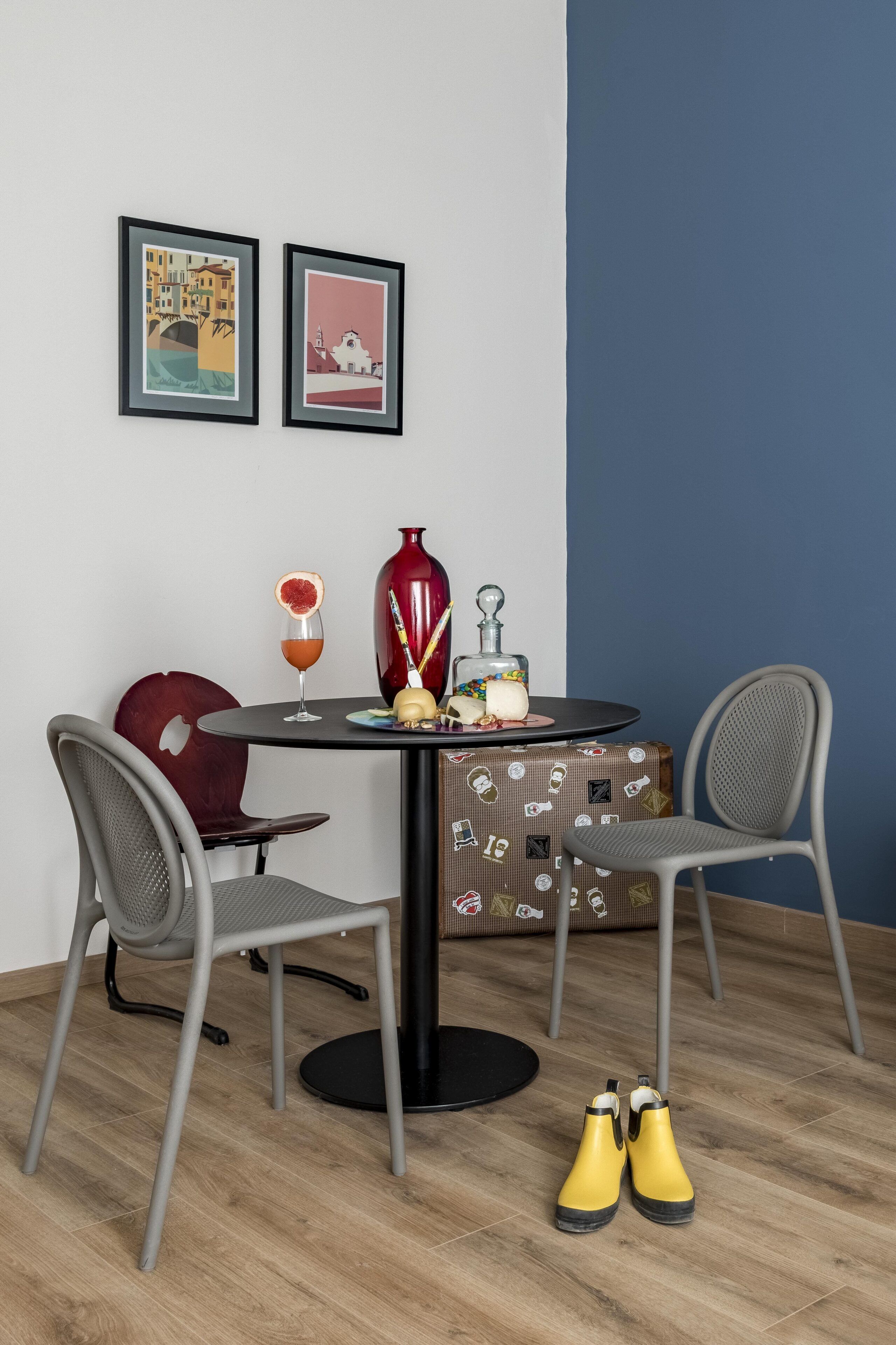 A dining area with a round table, two chairs, a red vase, a glass with a drink, and a cake.