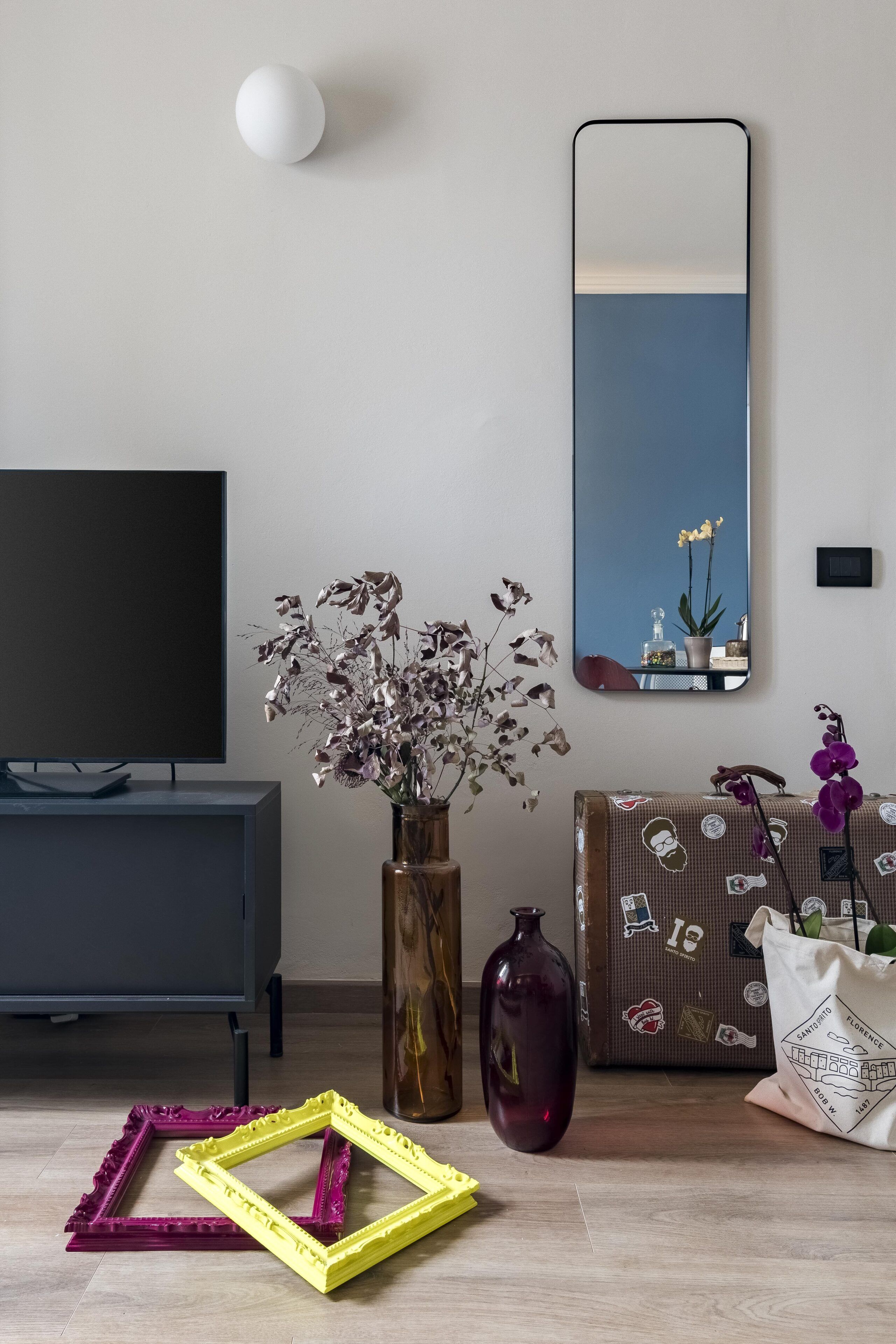 A modern living room with a flat-screen TV, a large mirror, a vase with dried flowers, a purple vase, and a suitcase with stickers.