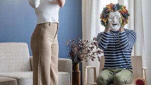 Twee personen in een kamer met een bank, een tafel met een drankje en een vaas met bloemen.