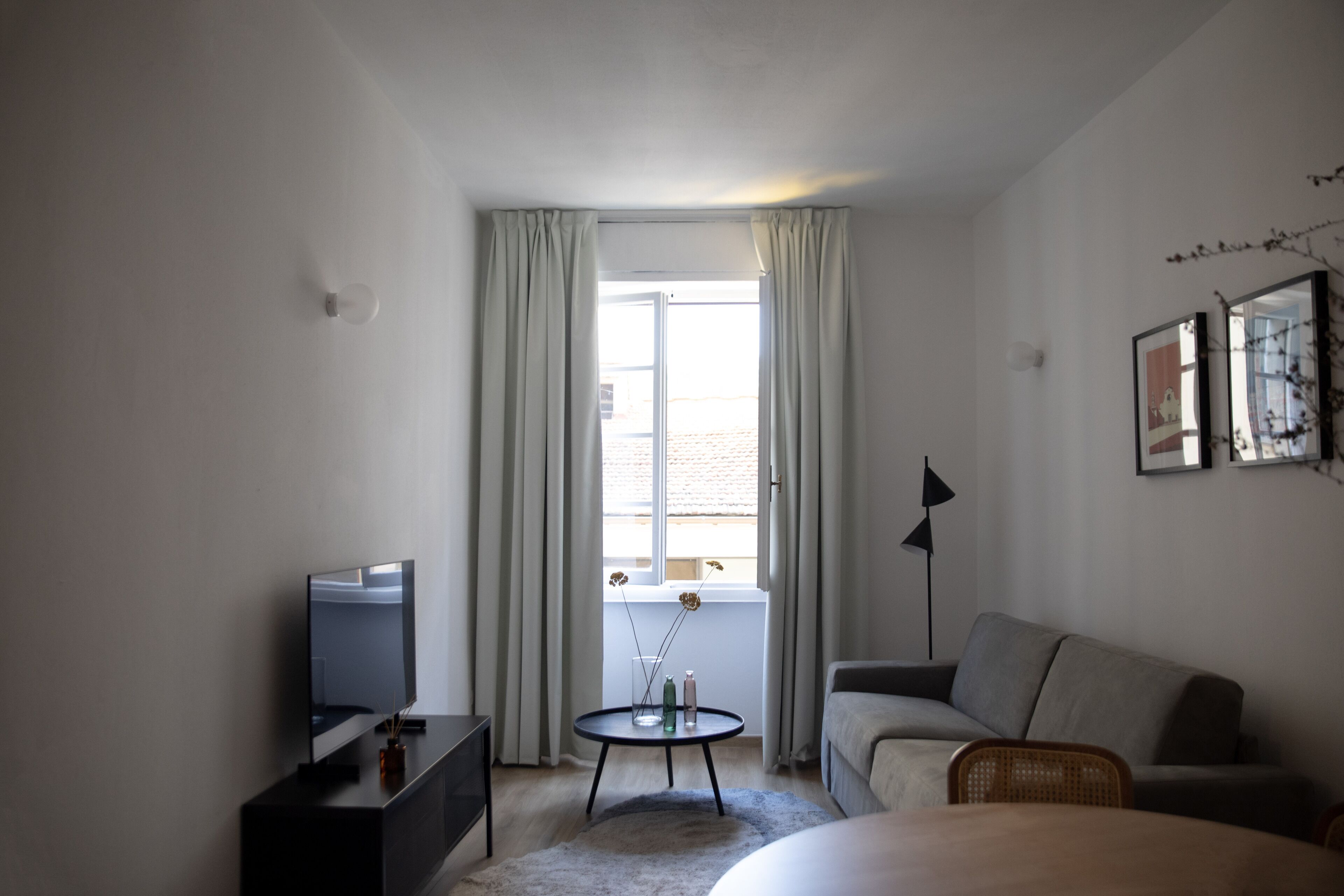 A modern hotel room with a bed, a sofa, a TV, a small table with decorative items, and a window with curtains.