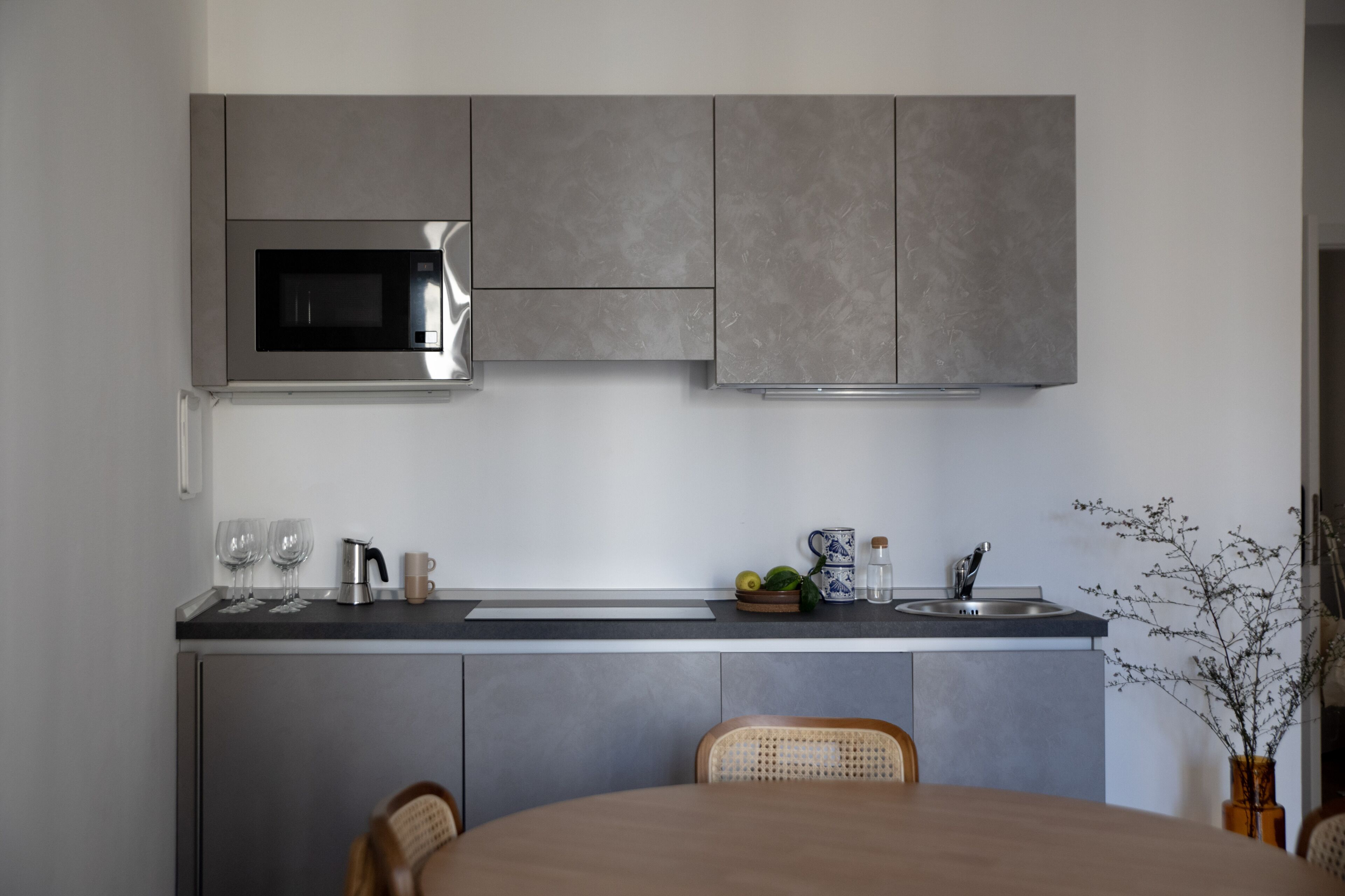 Una cucina moderna con mobili grigi, un forno a microonde in acciaio inossidabile e un lavello. In primo piano, due sedie e un tavolo da pranzo.