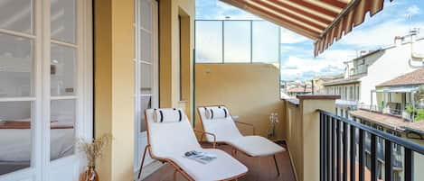 A balcony with two white lounge chairs, a small table, and a vase with dried plants.