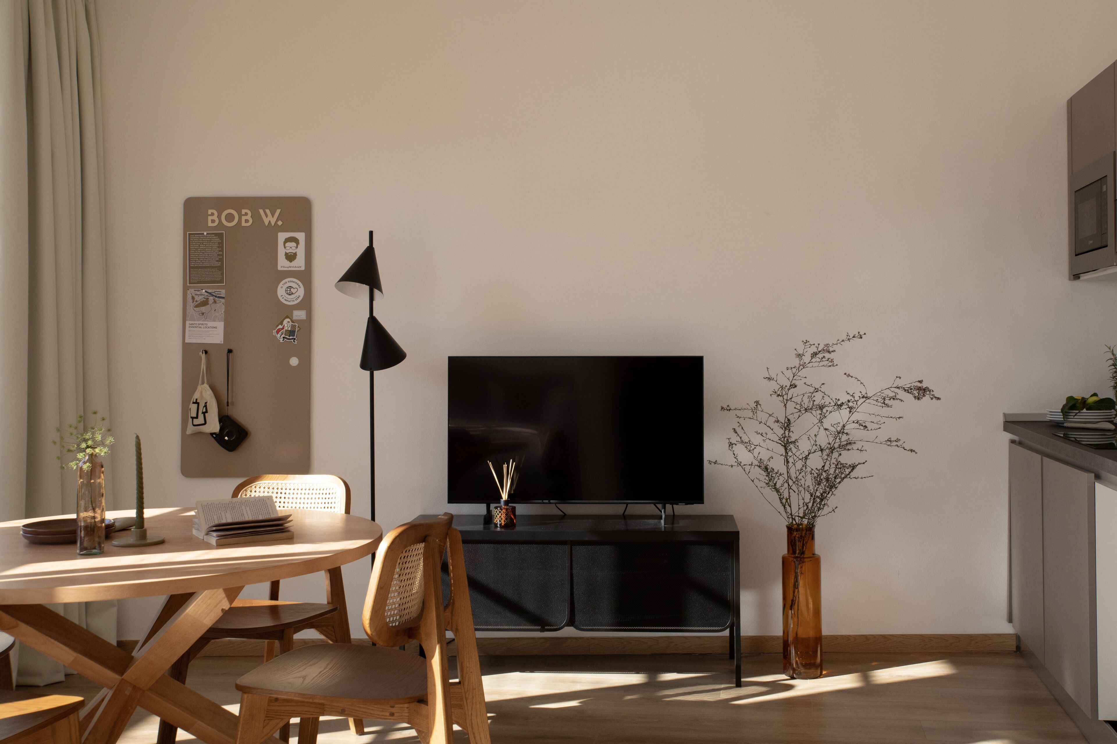 A modern living room with a wooden dining table, a black TV on a cabinet, a black floor lamp, and a vase with branches.
